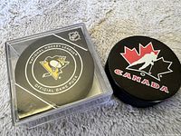 Photo showing the NHL Official Game Puck with Pittsburgh Penguins logo sealed in a clear protective case next to the Canada hockey puck