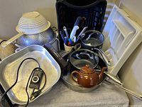 Photo showing kitchen items including strainers, pots, frying pan with lid, brown ceramic tea pot, electric fryer or warming tray, and white plastic utensil trays.