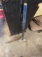 Sledge hammer standing vertically resting against a black object, blue handle visible, rusty pick axe head on the floor beside it.