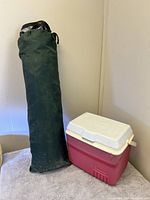 Green fabric tent packed in black cylindrical carrying bag with handles next to red plastic cooler with white lid on carpeted floor