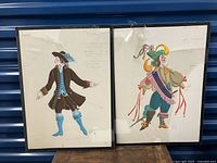 Two framed costume drawings displayed side by side, showing full view of both artworks