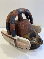 Front and angled side view of the carved wood mask showing the facial details, painted natural colors with the arch structure on top and white angular ear-like extensions on sides.