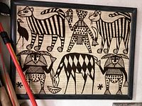 Framed African textile artwork depicting stylized animals and human figures in black designs on beige fabric, displayed vertically.