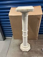 Wide shot of the full antique white marble column pedestal showing the octagonal base and square top shelf in front of a cardboard box and blue storage unit background.