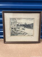 Framed lithograph artwork depicting a coastal scene with a dock, boats, buildings, and rocks in black and white.