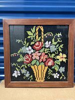 Framed folk art needlepoint showing a colorful floral basket embroidered on black fabric.