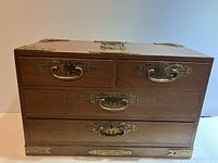 Front view of the wooden jewelry chest showing two small top drawers, one medium middle drawer, two large bottom drawers with ornate brass handles and decorative brass accents on the corners and top surface.