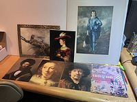 Photo of seven framed and unframed art prints on desk, showing various classical portraits and other subjects including a hunting bird and an Asian-inspired scene.