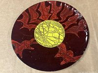 Front view of a round enamel plate showing abstract leaf-like design in red and brown with a yellow circular centerpiece on dark maroon background.