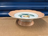 Front and side view of the peach-colored Mottahedeh cake stand with fruit design on the top surface on a wooden table with blue background.