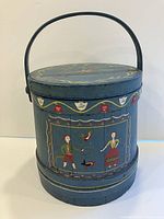 Front view of old blue wooden firkin with hand painted folk art figures of man, woman, and birds and a metal handle on top.