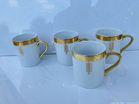 Four white ceramic mugs with gold bands and handles arranged on a white surface.