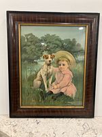 Framed lithograph showing a boy in pink attire sitting on grass next to a dog by a pond with lush greenery in the background. The frame is a faux bois original with wood grain look and gold inner border.