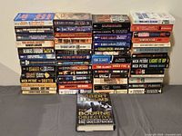 Photo showing arranged stack of approximately 37 books including authors Robert Ludlum, Tom Clancy, Nick Petrie, Stephen Coonts and others.