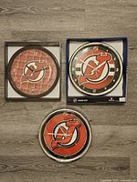 Three New Jersey Devils wall clocks arranged on the floor: one black framed without packaging, one chrome framed in box, and one chrome framed without packaging.