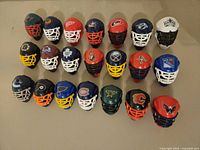 Photo showing all 21 mini plastic NHL goalie masks arranged in rows on a beige surface, clearly displaying various team colors and logos.