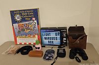 Full lot showing USA State Coin Collector's Map (new/sealed), Tzumi LED Light Up Marquee Box, men's black belt, binoculars with case, selfie stick, and small accessories arranged on table.