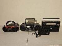 Four radios displayed side by side on a white surface against beige wall, showing the Realistic receiver, Soundesign boombox, Blackweb boombox, and Aiwa portable radio.