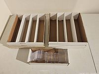 Two large white cardboard trading card storage boxes with four internal sections each, placed side by side. In front of them, a small white cardboard box containing assorted clear plastic trading card cases of various sizes, some hinged.