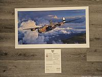 Full top view of the art print lying flat on floor with documentation centered below it. Shows entire artwork, aircraft flying over clouds.