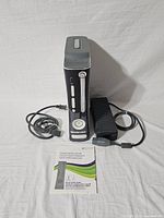 Xbox 360 console standing vertically with power supply unit and power cord coiled next to it. Quick setup guide booklet placed in front.