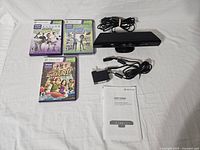 Shows three Xbox 360 Kinect games and the Kinect sensor with cables and booklet laid out on a white surface.