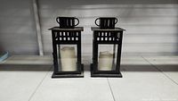 Two black metal candle lanterns displayed side by side, each containing a flameless white candle.