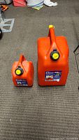 Two orange plastic gasoline jugs side by side - smaller 5 liter on left, larger 25 liter on right. Both have black and yellow screw caps with safety features.
