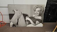 Large black and white photo of Marilyn Monroe reclining and smiling on the floor, wearing checkered pants and off-shoulder top, with her name text