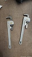 Photo showing both pipe wrenches side by side on carpet. Larger wrench on right, smaller on left. Metal wrenches with adjustable jaws and ridged gripping ends.