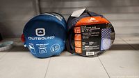 Two sleeping bags side by side in compression sacks, one blue labeled Outbound, other gray labeled Ozark Trail.