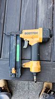 Full view of the Bostitch pneumatic nail gun showing yellow body, black handle, metal magazine, and air hose connector.