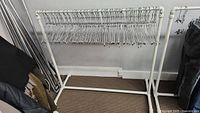 White metal clothes rack with about 100 chrome hangers hanging evenly on the horizontal bar, positioned against a wall on carpet flooring.