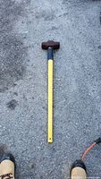 Full view of sledge hammer placed on gravel ground showing the yellow handle and rusted metal head.