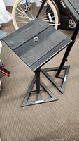 Photo of one black metal speaker stand showing square platform top and triangular base with a bicycle in the background.
