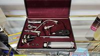 Top-down view of vintage medical diagnostic set items arranged in burgundy velvet-lined case showing otoscope, ophthalmoscope, tuning fork, specula, and handle.