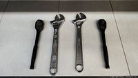 Four tools laid out on a white surface: two black Stanley ratchets and two adjustable wrenches with metal finish, some wear and slight rust on one wrench.