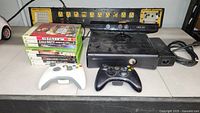 Xbox 360 console setup with two wireless controllers, Kinect camera, power cable, and nine game discs stacked on a shelf.