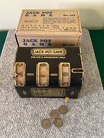 Photo showing the vintage Jack Pot Game slot machine with original box behind it on a surface. Four original coins placed in front of the machine. The game is black with gold detailing and has three display windows.