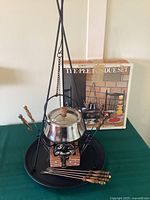Full view of the fondue set assembled with pot hanging from the tripod stand, 8 forks arranged around the base, and the original box in the background.