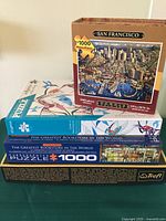 Photo shows the four puzzle boxes stacked: San Francisco folk art puzzle on top, Garden of Hummingbirds below, and two 'The Greatest Bookstore in the World' puzzles at the bottom. Each box states 1000 pieces.