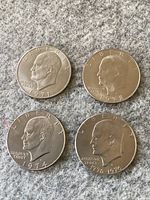 Four Eisenhower dollar coins dated 1971, 1972, 1974, and 1976 laid out on a grey fabric background, obverse side showing profile likeness of Dwight D. Eisenhower with inscriptions LIBERTY and the coin year.