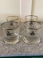 Four clear whiskey glasses with silver rims, featuring printed black image and text of The Nonsuch ship and Hudson's Bay Wine & Spirits Limited.