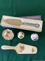 Top view of assorted English china pieces on green background including boxed Wedgwood cake server with blue floral handle, Royal Tudor Ware cake slice, Royal Albert Country Roses salt shaker and sugar bowl, and Hammersley Victorian Violets salt shaker.