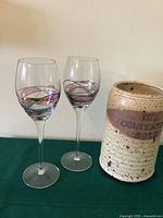 Two hand painted wine glasses with multi-color swirls placed next to a pottery wine chiller with speckled glaze and label.