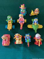 Nine colorful McDonalds Muppet Babies toys arranged on a green surface showing various characters on vehicles and ride-ons.