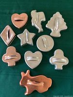 Top view of 10 vintage aluminum cookie cutters with handles, showing various shapes including heart, diamond, star, tree, circle, club, spade, and bunny. Four cutters are anodized aluminum with copper color.