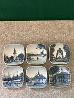 Six small square Royal Copenhagen plates each with unique blue and white location or architectural scene, laid out on carpet.
