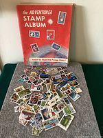 Pile of loose stamps in front of the The Adventurer Stamp Album, showing varied colorful stamps with different designs and subjects.