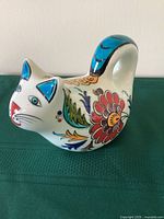 Front side view of ceramic cat figurine showing colorful floral and leaf motifs, turquoise tail, and facial details.